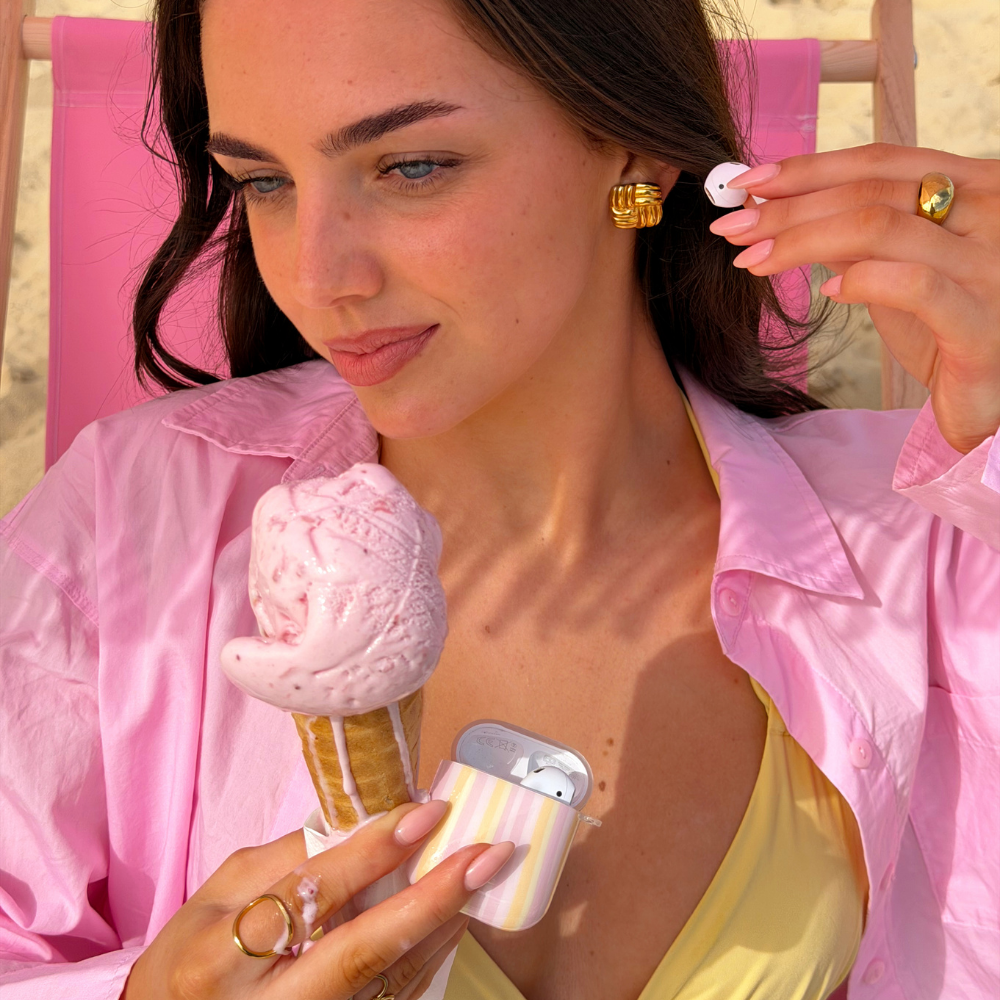 Girl putting airpods in her ear holding ice cream on the beach with her airpods case pastel stripes