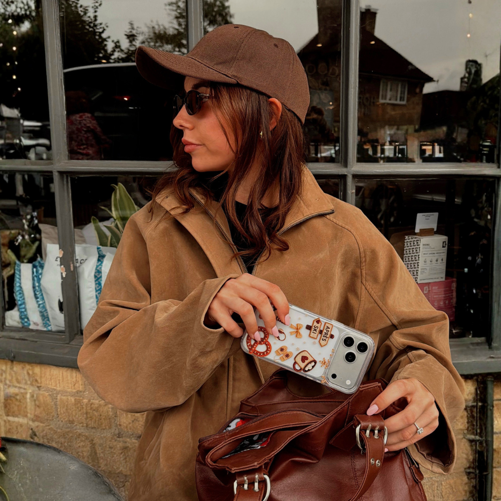 A woman wearing a brown suede-like jacket, sunglasses, and a hat sits at a cafe table with a croissant, coffee, and a phone case featuring winter themed illustrations including a coffee mug, ice skate, pretzel and a sign saying Apres Hour.