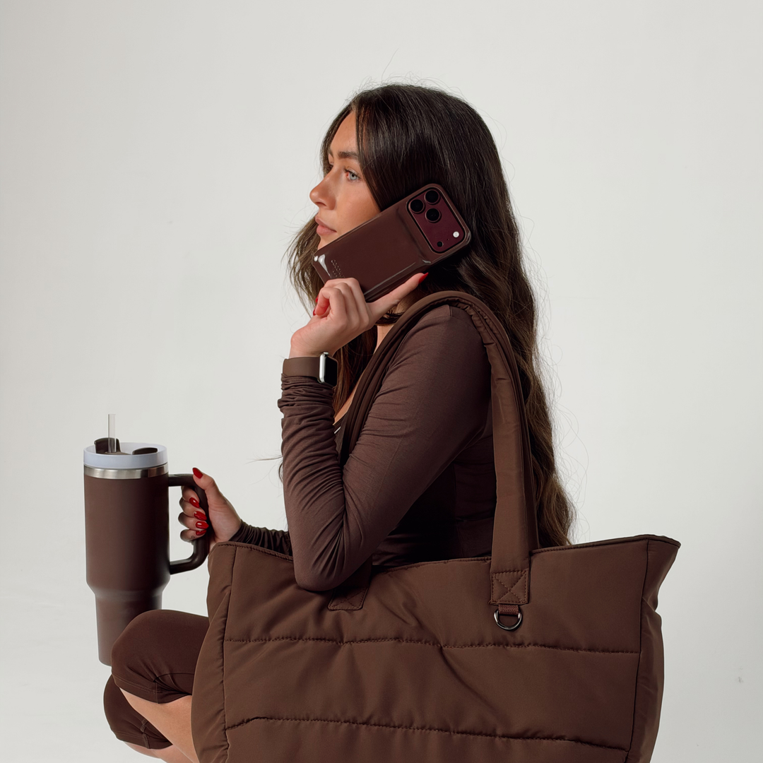 Woman wearing a brown pilates outfit, holding an iphone up to her ear. The phone is in a glossy chocolate brown Coconut Lane puffer phone case with embossed logo. She has The Main Carry Choc puffer bag on her shoulder and is holding the Chocolate brown tumbler. On her wrist is a Coconut Lane NAKD Chocolate brown apple watch strap.