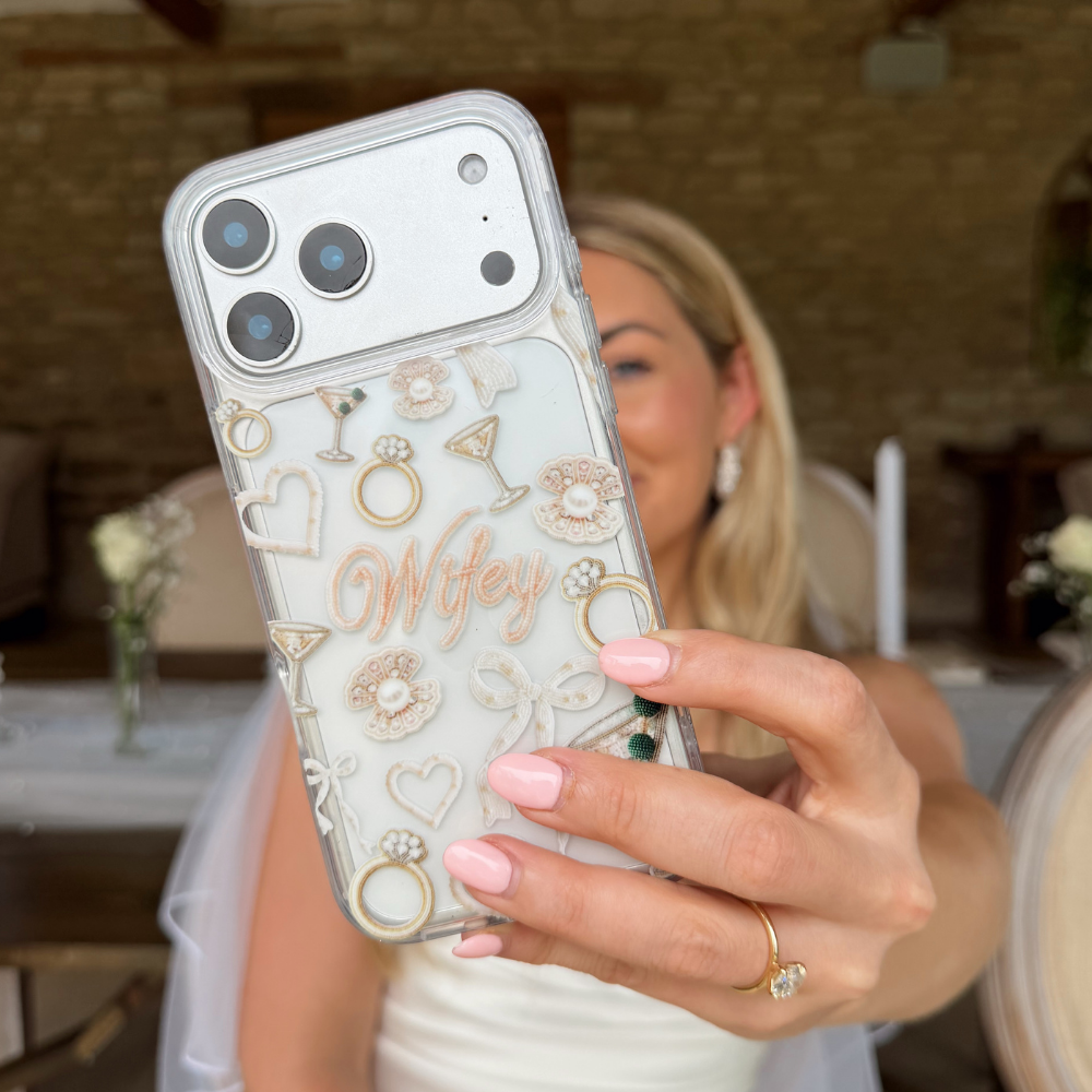 Bride holding a phone case with rings, hearts, cocktails, bows, pearls and the word 'Wifey'