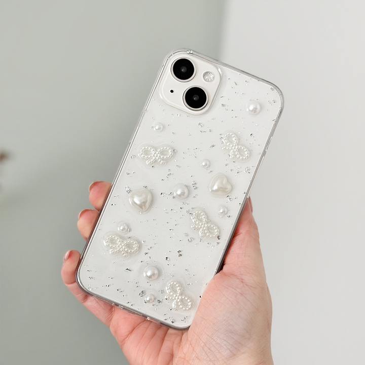 A hand holds an iPhone15 with a Coconut Lane Coquette Dreams Phone Case. The decorative case features embedded pearls, butterfly shapes, and sparkling embellishments on a translucent background, offering extra protective qualities. The phone has dual cameras and a sleek, glossy finish. The background is blurred.