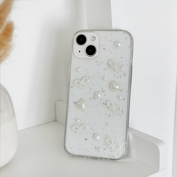A silver smartphone adorned with the Coquette Dreams Phone Case from Coconut Lane, featuring elegant pearls and bead designs, stands upright on a white shelf. In the background, a vase filled with beige dried grasses adds to the serene setting. The iPhone15 Case boasts two rear cameras, enhancing the minimalistic and stylish arrangement.