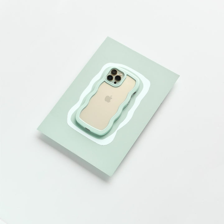 A beige iPhone 15 with a triple camera setup is encased in a Curvy Phone Case - Mint by Coconut Lane. It is placed on a light green rectangular surface, against a clean, white background.