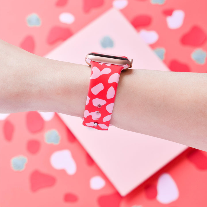 A person's wrist adorned with a Coconut Lane Red & Pink Leopard Apple Watch Strap, showcasing an eye-catching animal print on the silicone band. The backdrop echoes the theme with a matching red and pink abstract pattern, achieving a cohesive and stylish appearance.