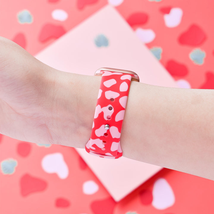 A close-up shot showcases a wrist adorned with the Coconut Lane Red & Pink Leopard Apple Watch Strap. The backdrop echoes the strap's design with abstract red, pink, and white shapes. This silicone band delivers a bold, cohesive look with its striking animal print pattern.