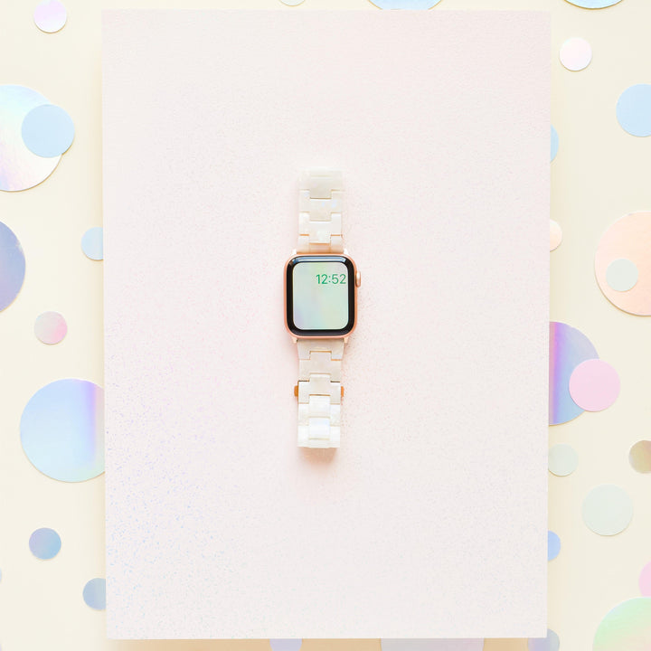 A smartwatch featuring the Luxe Iridescent Apple Watch Strap from Coconut Lane, with a white ceramic band, is displayed against a pastel background with holographic dots. The watch screen shows the time as 12:52. The minimalist and modern design perfectly complements the stylish and chic setup, further enhanced by hints of the iridescent watch strap.