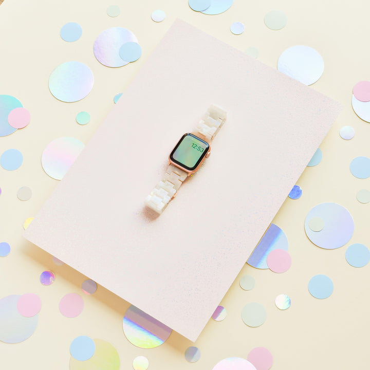 A Luxe Iridescent Apple Watch Strap by Coconut Lane, featuring a white band, is displayed on a pastel pink rectangular surface. Surrounding the strap and surface are variously sized, iridescent circular confetti pieces scattered across a light beige background, perfectly highlighting its sleek appearance.