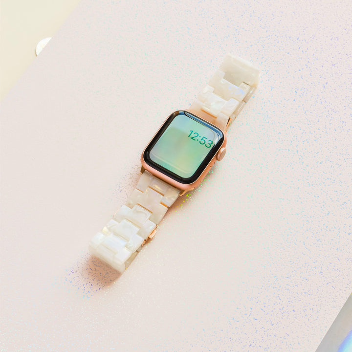 A Luxe Iridescent Apple Watch Strap by Coconut Lane, featuring a rectangular screen displaying the time "12:53", is shown on a light pink, slightly textured surface. The watch boasts a rose gold frame and an iridescent watch strap with a glossy white marbled pattern.