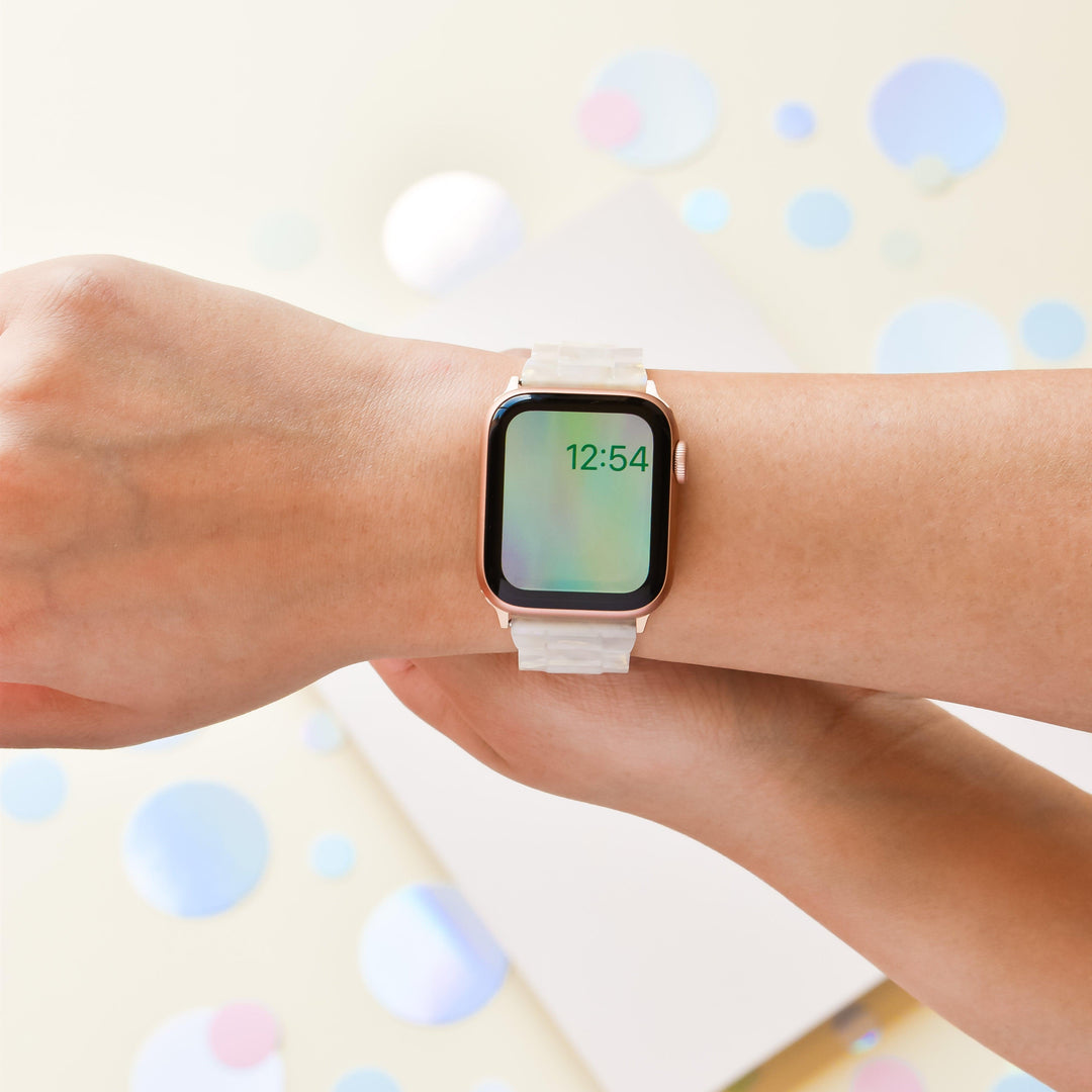 A person wearing a Luxe Iridescent Apple Watch Strap from Coconut Lane, featuring a rectangular face displaying the time, 12:54. The background is decorated with scattered pastel-colored circles and a white rectangular object. The person's arm is positioned to show the watch clearly.