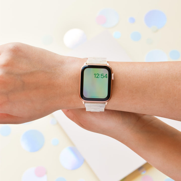 A person wearing a Luxe Iridescent Apple Watch Strap from Coconut Lane, featuring a rectangular face displaying the time, 12:54. The background is decorated with scattered pastel-colored circles and a white rectangular object. The person's arm is positioned to show the watch clearly.