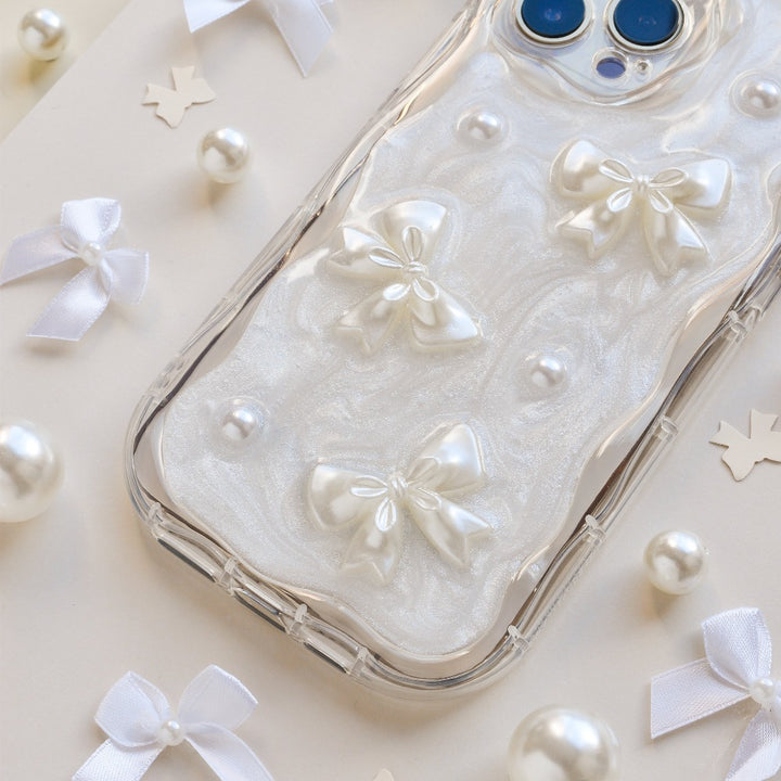 A smartphone in a Coconut Lane Pearls & Bows Phone Case, adorned with pearlescent bows and pearls, rests on a light surface scattered with white ribbons and small pearls. The phone's dual-camera lenses are visible at the top right. The image exudes a soft, elegant aesthetic.