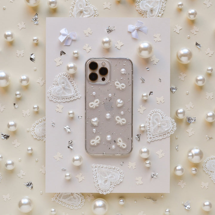 A Coquette Dreams Phone Case by Coconut Lane for the iPhone 15 lies in the center of a beige background adorned with pearls, lace hearts, and tiny bows. The intricate details of this extra protective case and the surrounding decorations exude a delicate, elegant aesthetic.