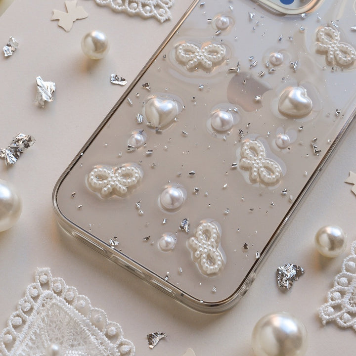 A close-up image of the Coquette Dreams Phone Case by Coconut Lane, a transparent iPhone 15 case adorned with 3D white butterfly and heart designs, as well as pearl-like embellishments. Extra protective, the phone rests on a white surface surrounded by decorative pearls, lace cutouts, and silver confetti pieces.