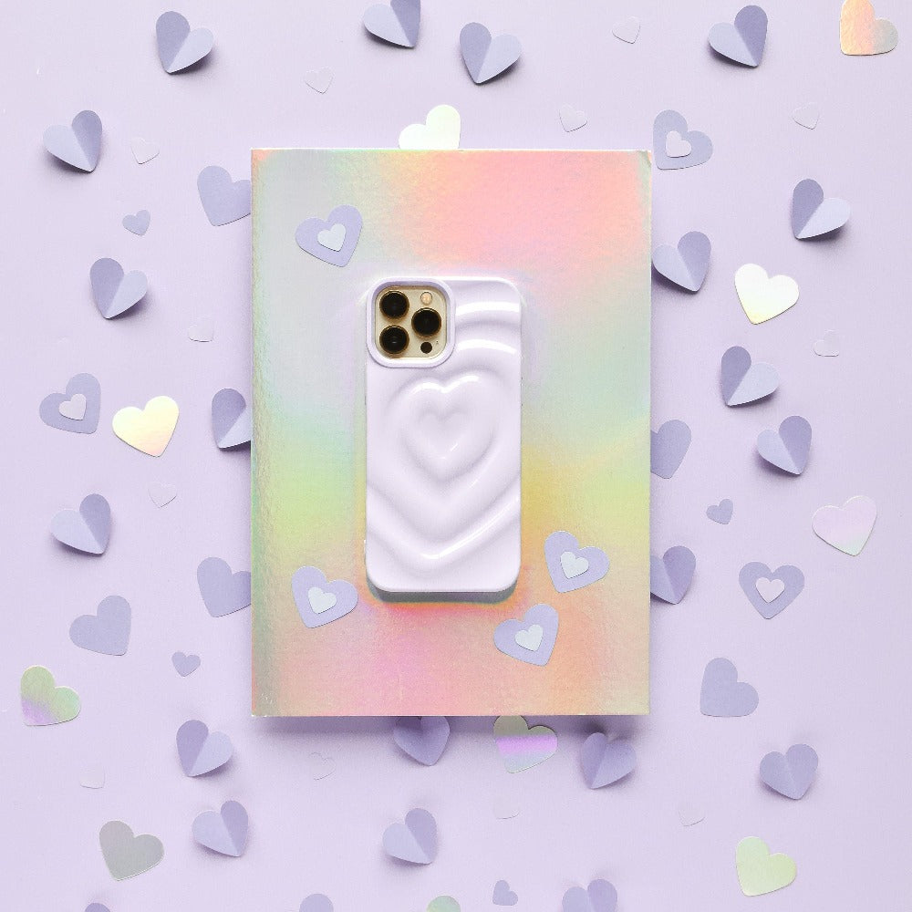 A smartphone adorned with Coconut Lane's Melting Heart Phone Case in Lilac is resting atop an iridescent, pastel-colored notebook. The background features a shimmering lilac hue, scattered with small cut-out paper hearts in shades of lavender and silver.