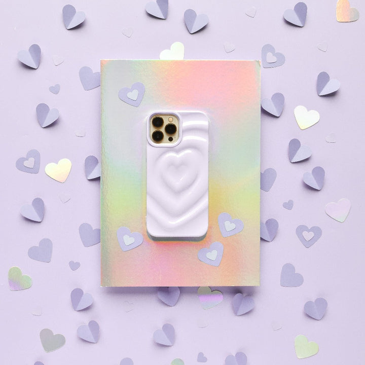 A smartphone adorned with Coconut Lane's Melting Heart Phone Case in Lilac is resting atop an iridescent, pastel-colored notebook. The background features a shimmering lilac hue, scattered with small cut-out paper hearts in shades of lavender and silver.