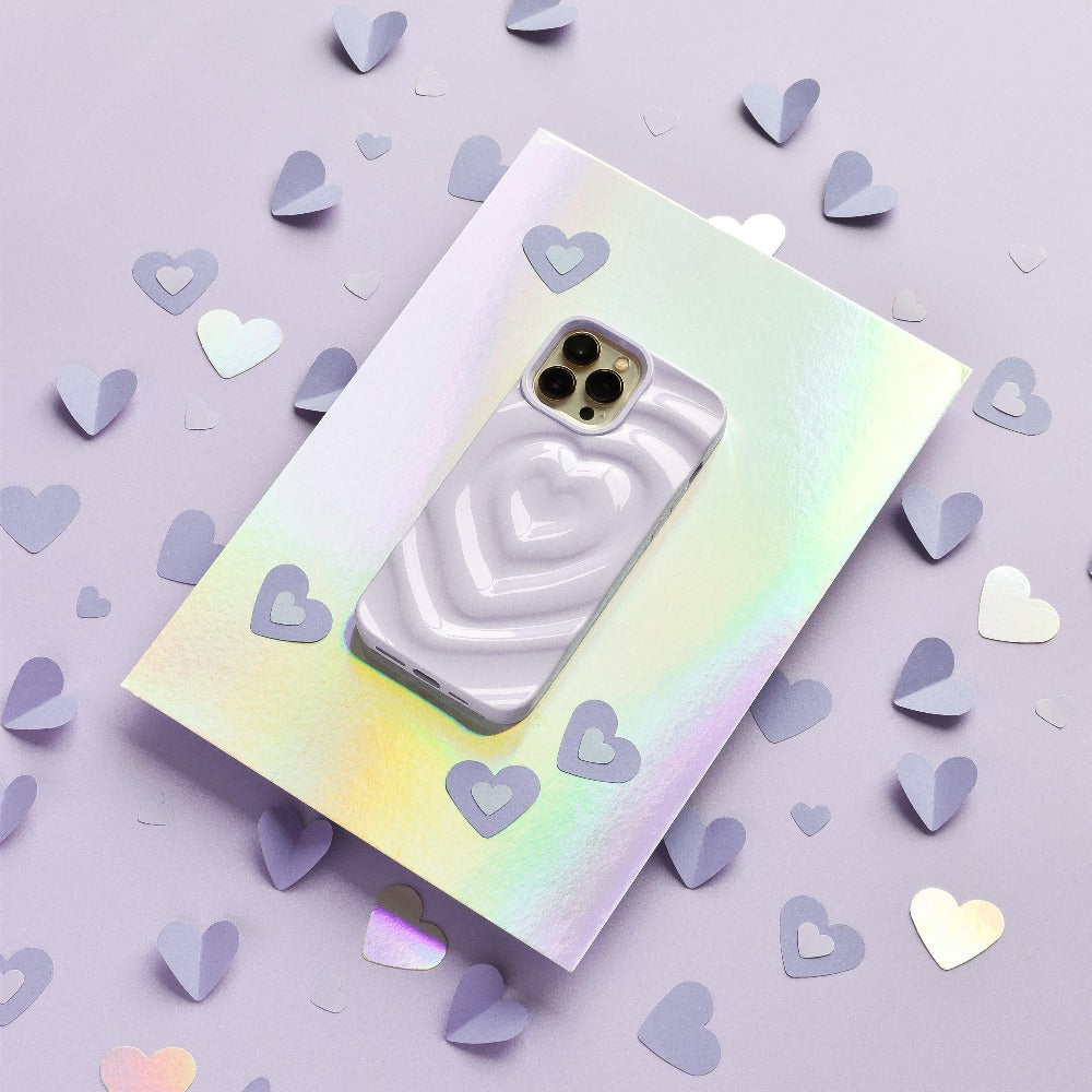 A smartphone with Coconut Lane's Melting Heart Phone Case in Lilac, featuring a textured 3D melting heart effect, rests on a holographic sheet. The background is a shimmering lilac surface adorned with scattered purple paper hearts of various sizes.