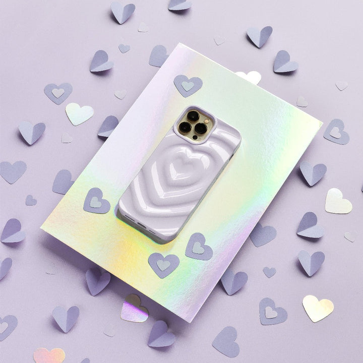 A smartphone with Coconut Lane's Melting Heart Phone Case in Lilac, featuring a textured 3D melting heart effect, rests on a holographic sheet. The background is a shimmering lilac surface adorned with scattered purple paper hearts of various sizes.