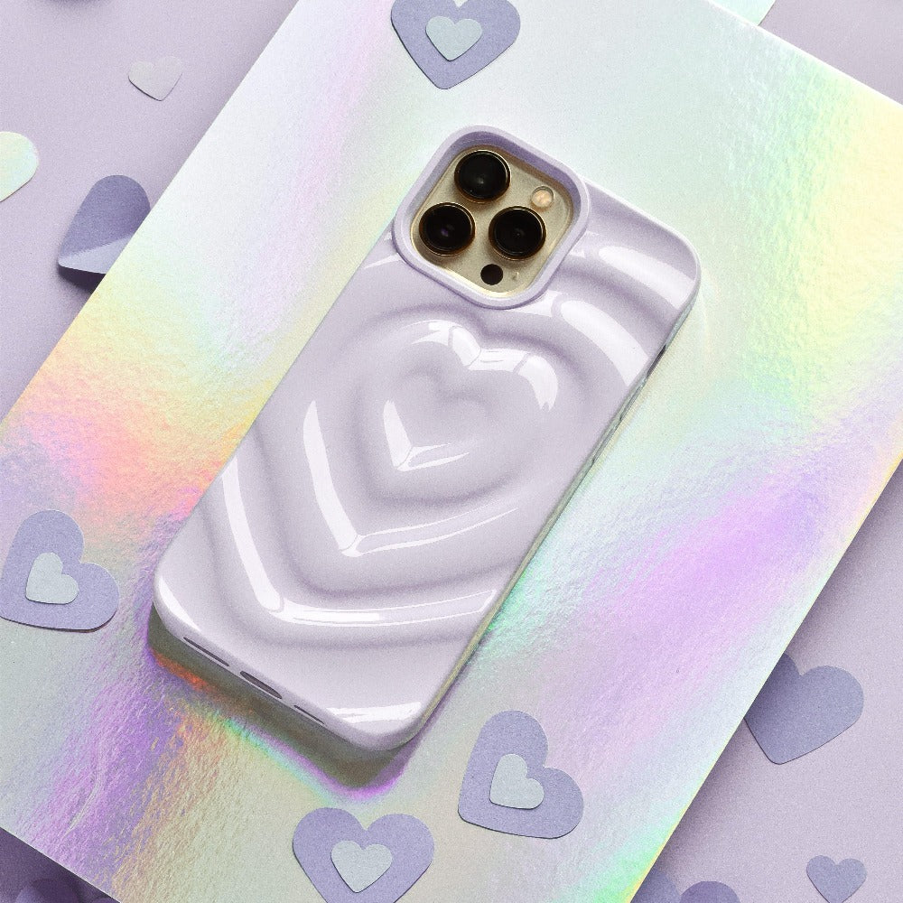 A shiny iPhone with a Melting Heart Phone Case in lilac from Coconut Lane, featuring a raised heart design, lies on a holographic surface. Surrounding the phone are small, flat, heart-shaped cutouts in matching lavender, adding to the overall whimsical and romantic aesthetic.