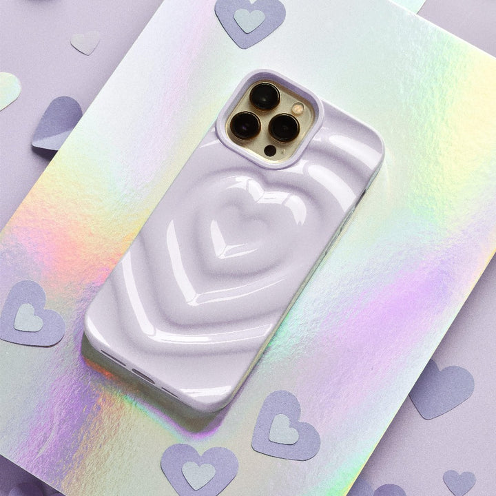 A shiny iPhone with a Melting Heart Phone Case in lilac from Coconut Lane, featuring a raised heart design, lies on a holographic surface. Surrounding the phone are small, flat, heart-shaped cutouts in matching lavender, adding to the overall whimsical and romantic aesthetic.