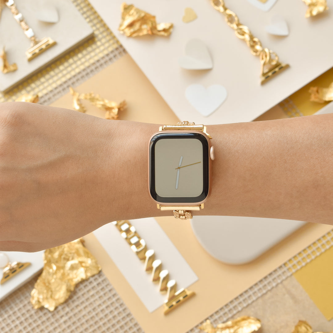 A person's wrist adorned with a gold smartwatch featuring the Square Gold Bracelet Apple Watch Strap by Coconut Lane is shown against a backdrop of gold and white decorative elements, including gold leaves, white hearts, and various textured materials. The environment exudes luxury and aesthetic appeal.