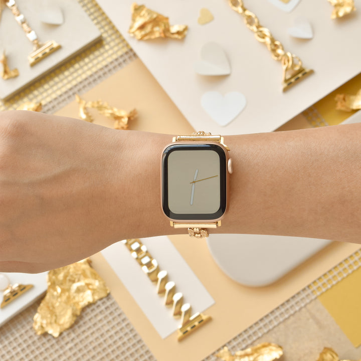 A person's wrist adorned with a gold smartwatch featuring the Square Gold Bracelet Apple Watch Strap by Coconut Lane is shown against a backdrop of gold and white decorative elements, including gold leaves, white hearts, and various textured materials. The environment exudes luxury and aesthetic appeal.