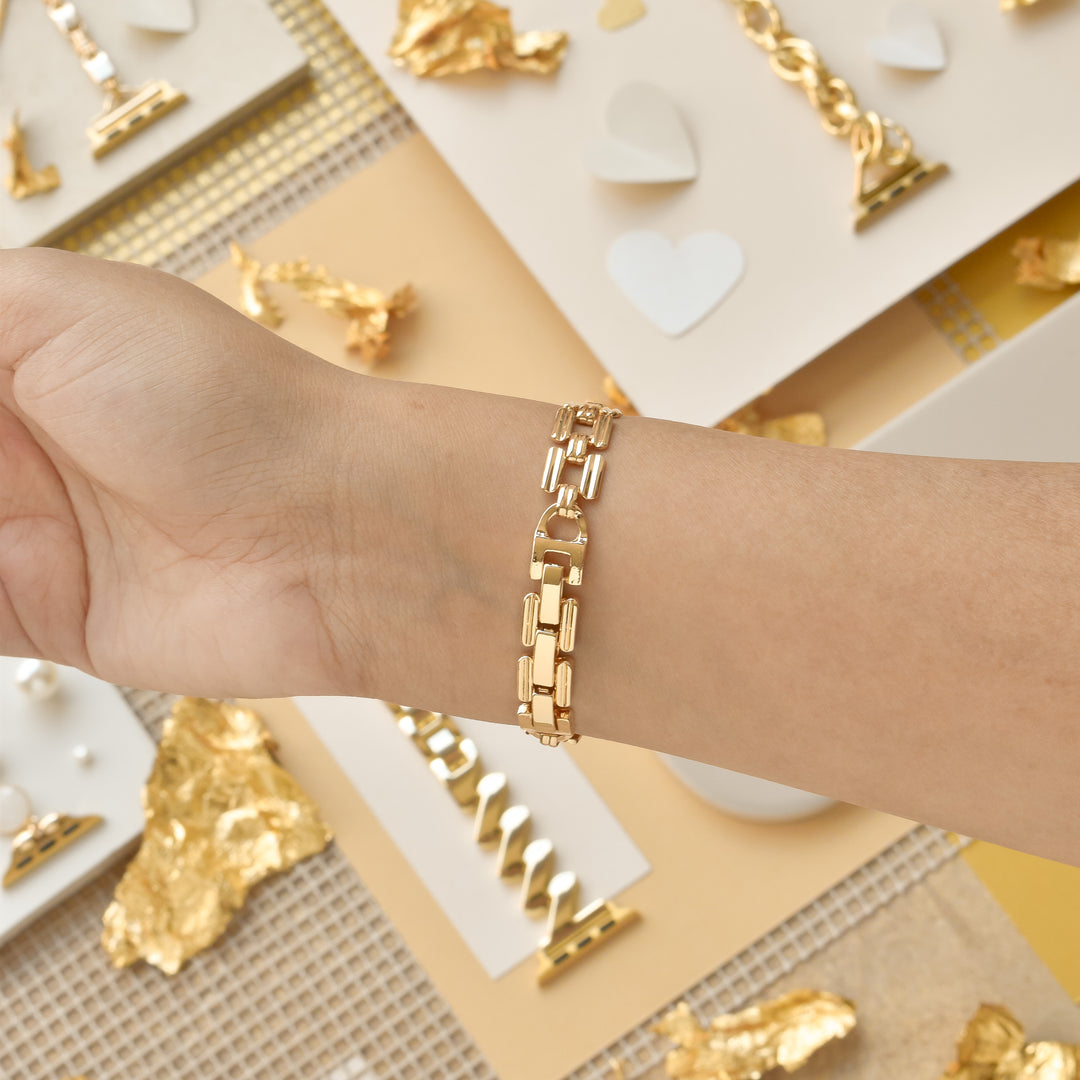 A close-up of a person wearing the Square Gold Bracelet Apple Watch Strap by Coconut Lane on their wrist. The background features a yellow and beige theme with gold foil accents, small hearts, and jewelry pieces like necklaces and rings displayed on white and beige surfaces.