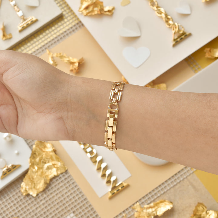 A close-up of a person wearing the Square Gold Bracelet Apple Watch Strap by Coconut Lane on their wrist. The background features a yellow and beige theme with gold foil accents, small hearts, and jewelry pieces like necklaces and rings displayed on white and beige surfaces.