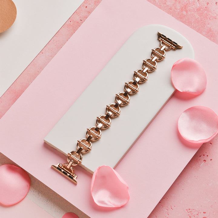 The Metal Link Apple Watch Strap - Rose Gold by Coconut Lane is showcased on a white, oval-shaped platform. The strap's interlocking circular links are complemented by scattered pink rose petals, all placed on a textured pink and white background, evoking an air of elegance.