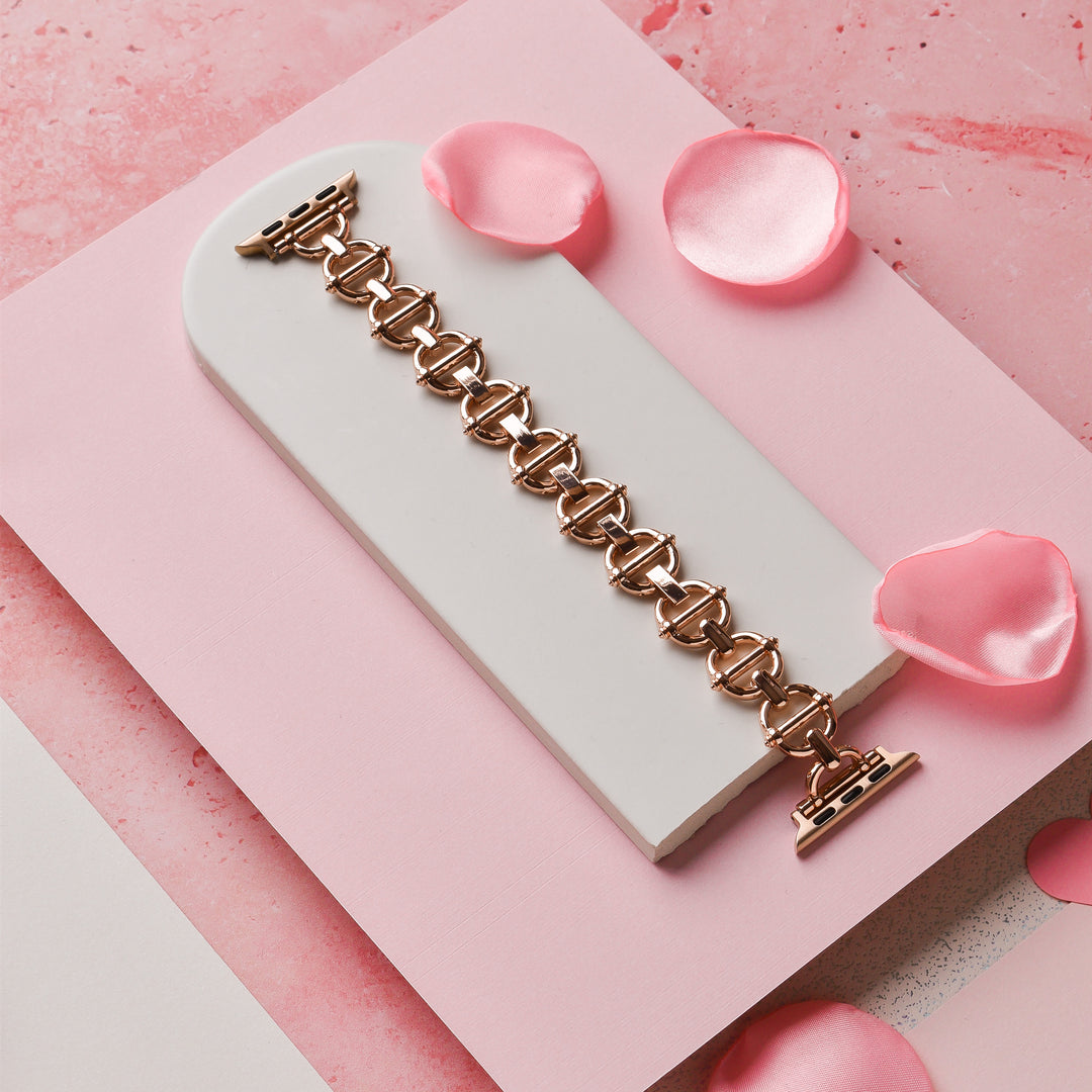 The Metal Link Apple Watch Strap in Rose Gold by Coconut Lane lies on a white rectangular surface, surrounded by scattered pink rose petals. The background features pink and white hues with a marbled texture.