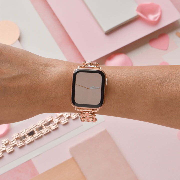 A close-up of a woman's arm wearing the Coconut Lane Metal Link Apple Watch Strap in Rose Gold. The watch face showcases a minimalistic design. The background features pink and peach-colored paper, a chain-link bracelet, and pink petals scattered around.