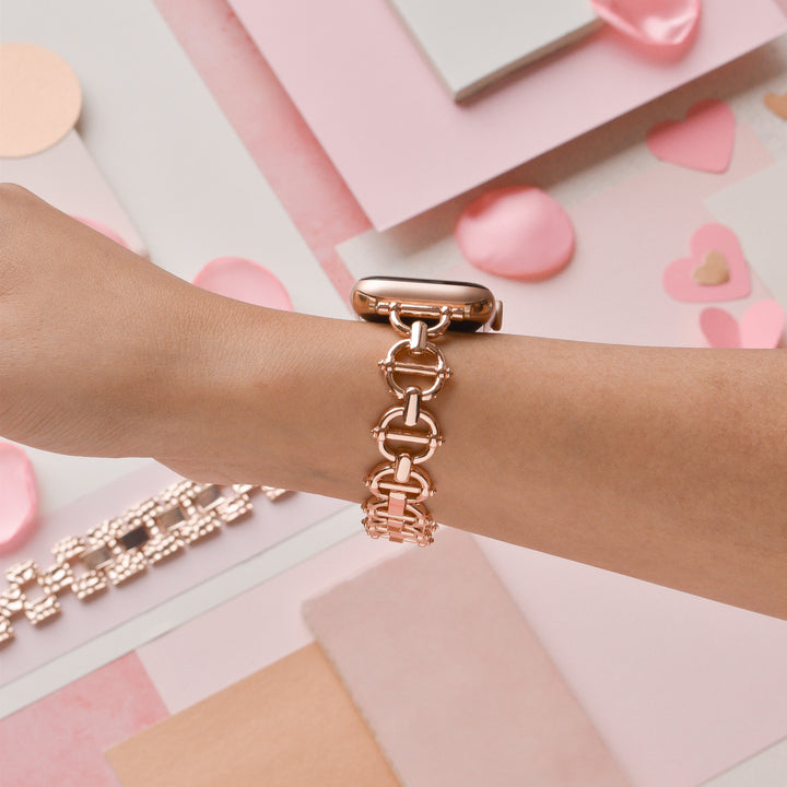 A person's wrist adorned with a stylish Coconut Lane Metal Link Apple Watch Strap in rose gold. The background features pastel-colored papers, heart-shaped decorations, and other accessories, giving a soft, romantic feel. The design beautifully complements the stainless steel elements of the strap.