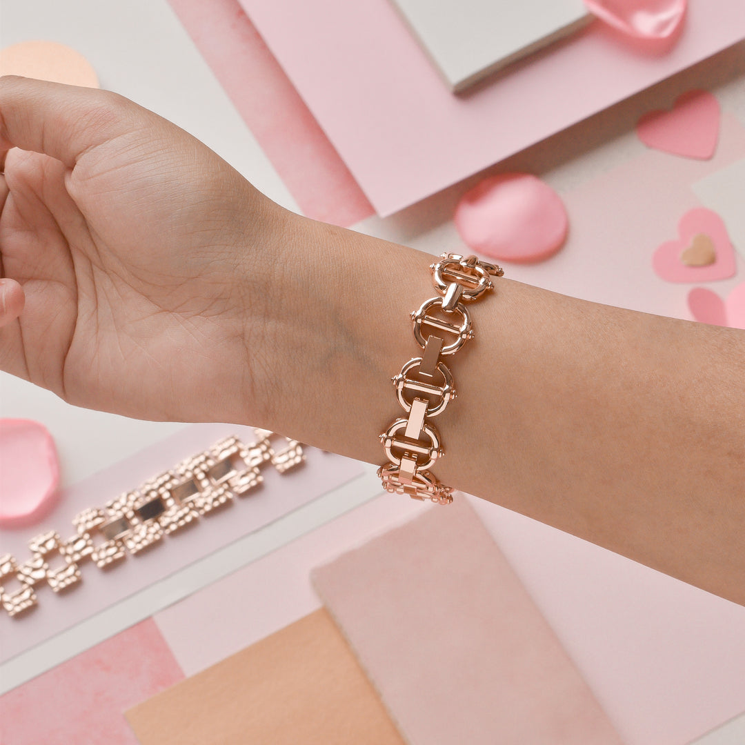 A person's hand wearing a gold bracelet with a chunky interlocking design is showcased. The background features pink, white, and beige-colored papers along with decorative heart shapes, enhancing the romantic and elegant theme. Beside the bracelet is Coconut Lane's stylish Metal Link Apple Watch Strap in Rose Gold, adding a touch of modern sophistication.