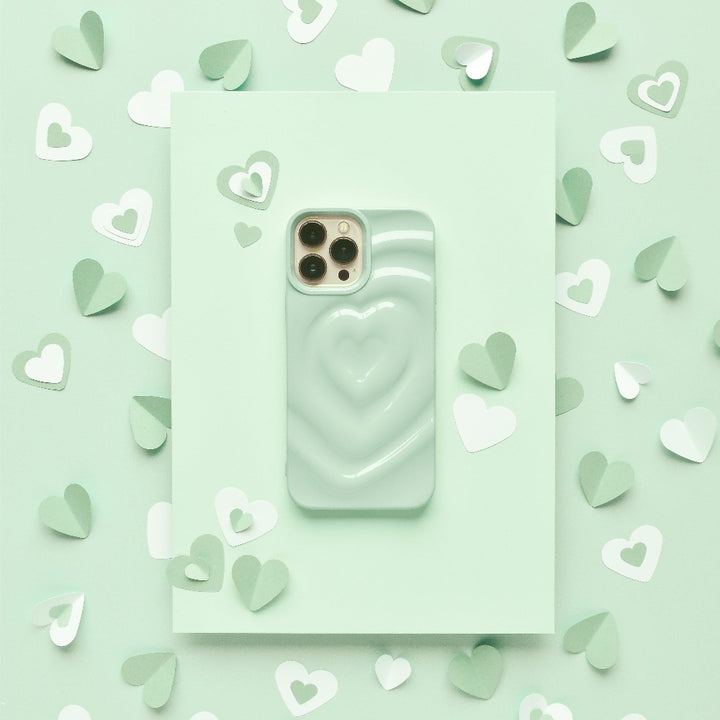 A smartphone with a Melting Heart Phone Case in Sage from Coconut Lane, featuring an extra protective raised heart design, is placed on a light green surface. Surrounding the phone are small paper hearts in various shades of green and white, creating a charming, romantic aesthetic.