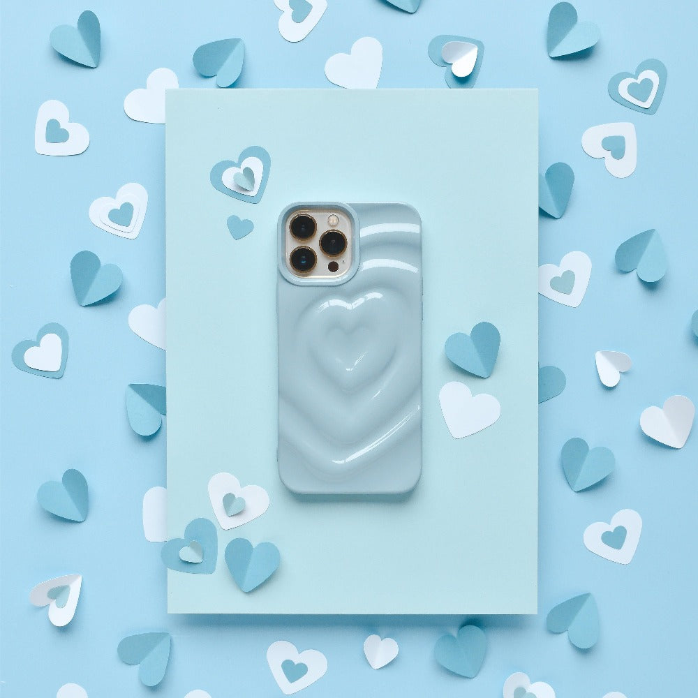 A smartphone encased in Coconut Lane's Melting Heart Phone Case in Baby Blue is positioned atop a larger light blue sheet of paper. Encircling the phone are numerous small heart-shaped cutouts in white and blue hues. The entire setting features a light blue background, establishing a harmonious color palette.