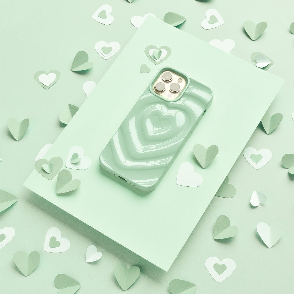 A smartphone encased in the Melting Heart Phone Case - Sage by Coconut Lane, featuring a sage green and white textured melting heart design, is placed on a green background. The setup includes paper hearts in various shades of green and white, offering a coordinated and visually appealing display.