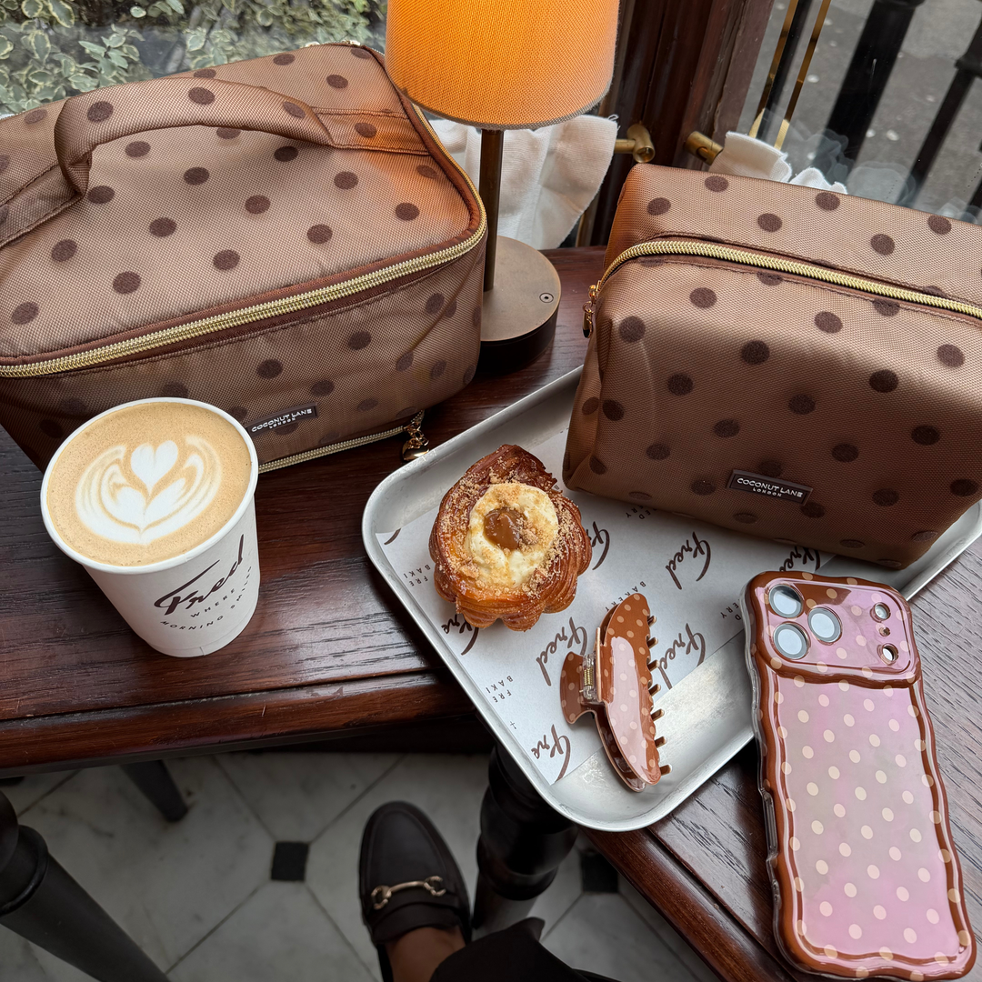 Flat lay cafe photo of the Dot Society chocolate brown collection. The vanity case is a brown mesh material with brown polka dot detailing. In the photo is the matching wavy phone case, claw clip and makeup bag.