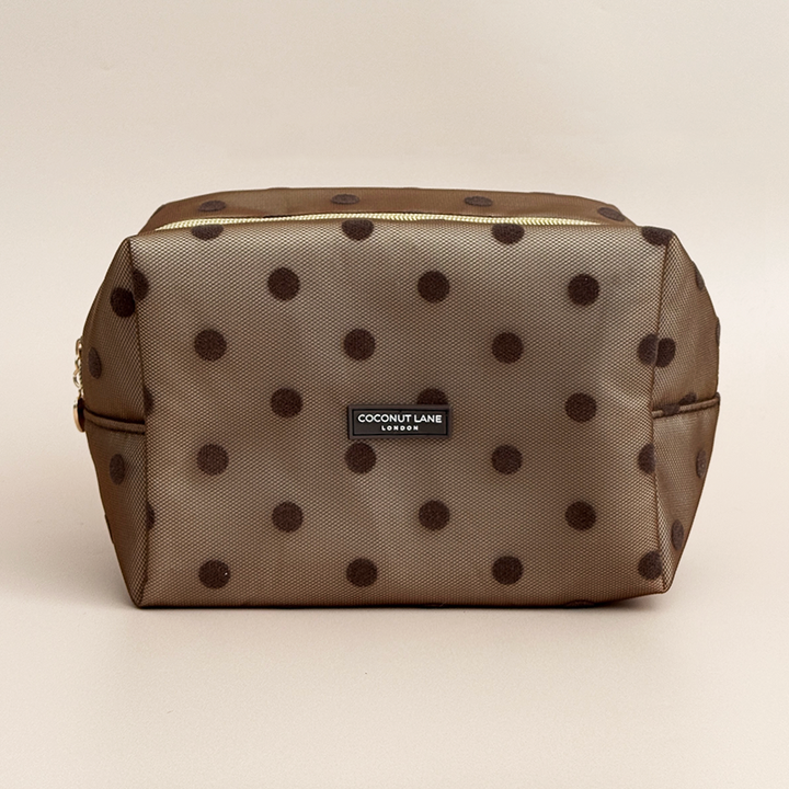 Brown mesh makeup bag with dark brown polka dots, a gold top zip with a heart-shaped zip pull, and a black Coconut Lane London logo badge on the front, shown on a beige background.
