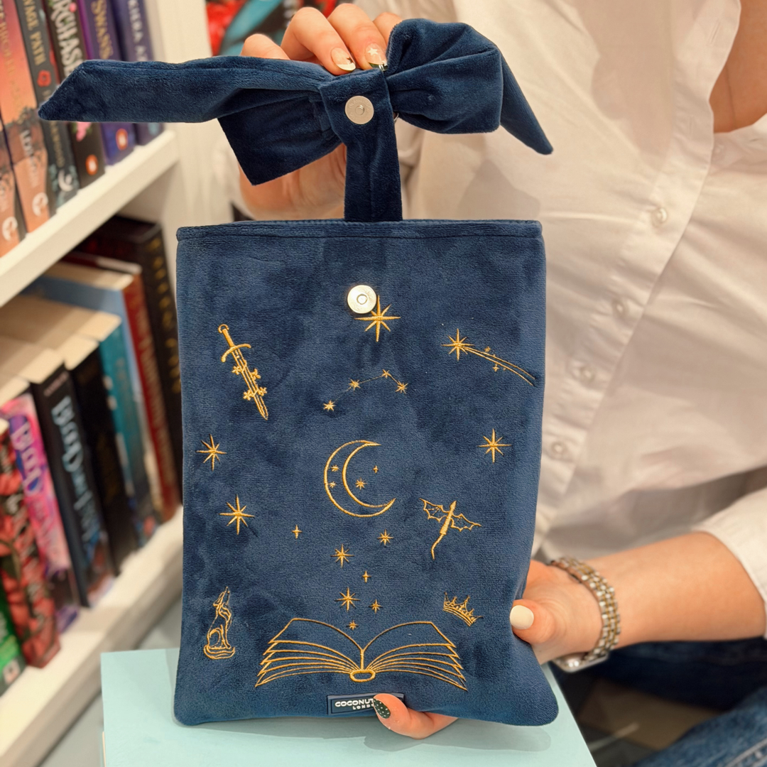 Book Sleeve made from soft navy blue velvet with gold embroidered crescent moon, stars, crown and open book illustrations, finished with a large velvet bow and Coconut Lane logo, photographed on a white background.