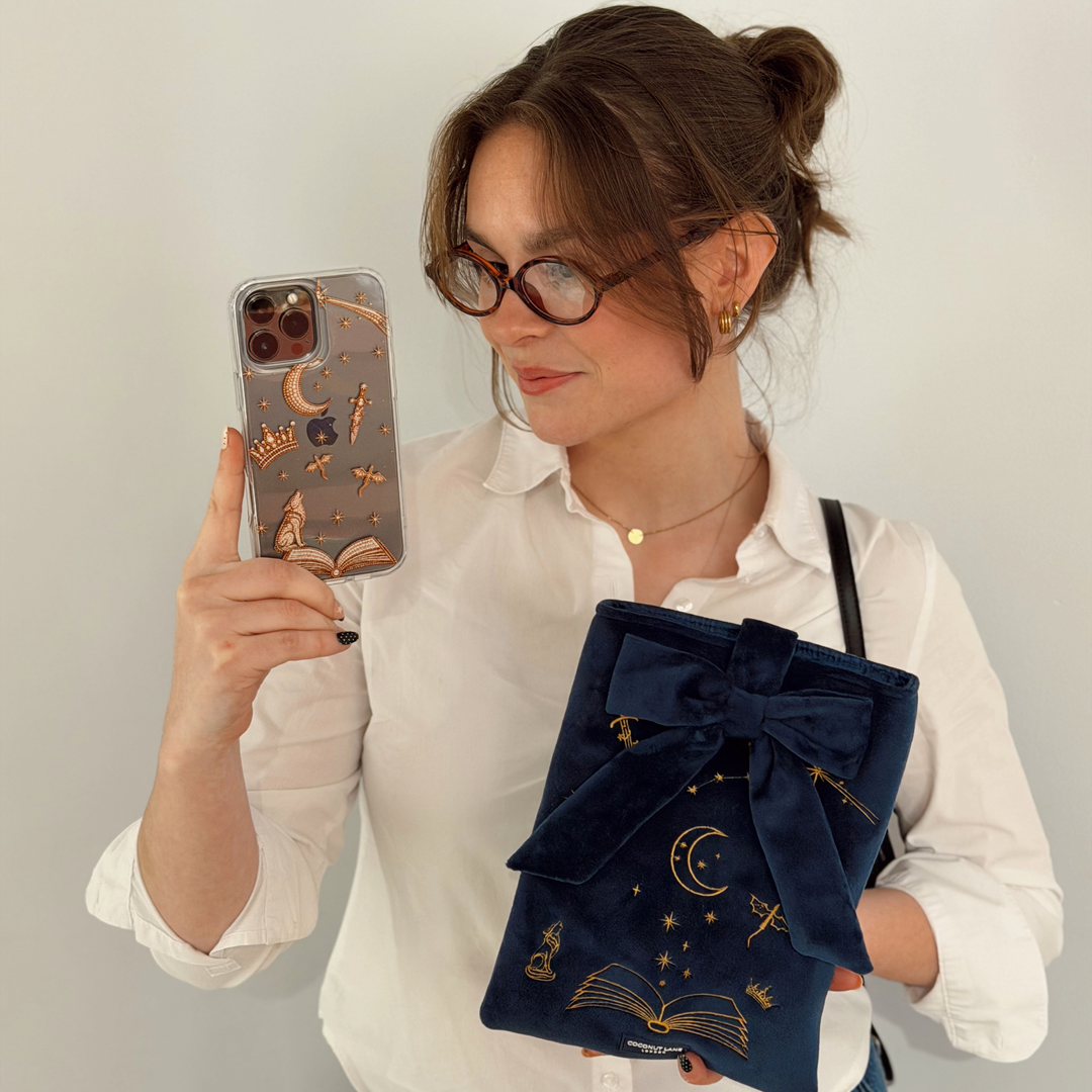 Book Sleeve made from soft navy blue velvet with gold embroidered crescent moon, stars, crown and open book illustrations, finished with a large velvet bow and Coconut Lane logo, photographed on a white background.