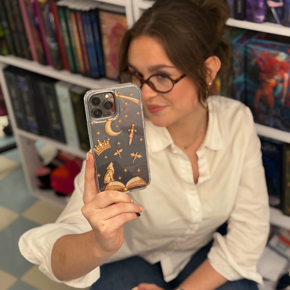 Woman posing taking a selfie, holding an iphone with a clear fantasy girl phone case with golden illustrations of fantasy themed beaded effect icons, such as a book, wolf, crown, moon, sword, stars and dragons.