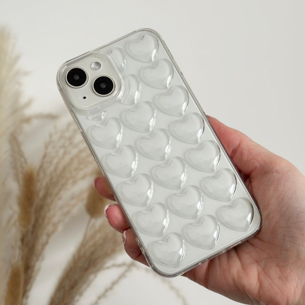 A hand holds an iPhone 15 in the Coconut Lane 3D Hearts Phone Case - Clear. The case features multiple raised, heart-shaped bubbles on the back. The background remains minimal, showcasing some blurred beige pampas grass to the left side.