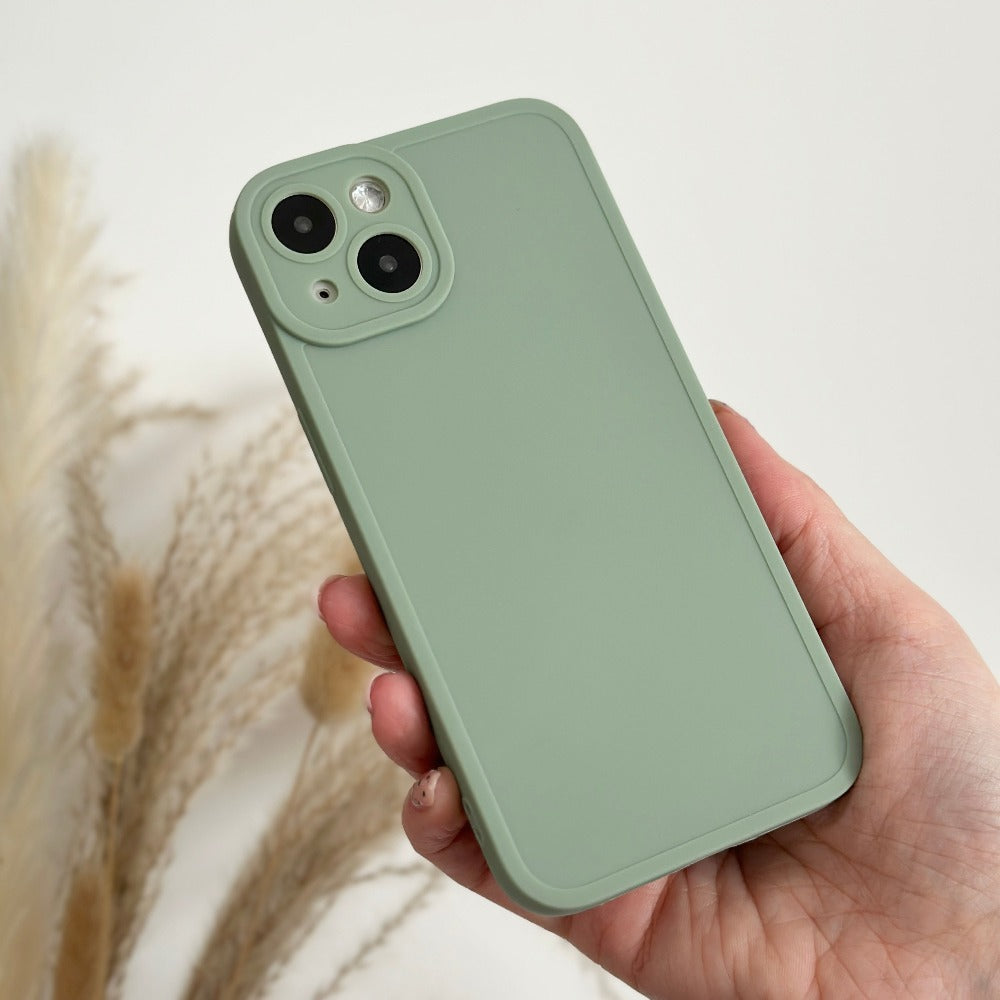 A hand holding a green Coconut Lane NAKD Phone Case in Sage, which has a plain, matte finish. The smartphone features dual cameras and an LED flash on the back, fully protected by the case. The background includes soft, beige pampas grass against a plain white wall.