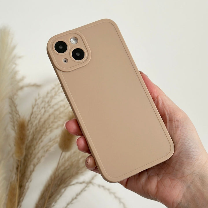 A hand is holding a beige smartphone encased in a Coconut Lane NAKD Phone Case - Nude, which offers full camera protection. The dual cameras and flash are visible in the upper left corner of the back. In the background, dried grasses are softly focused against a light-colored wall.