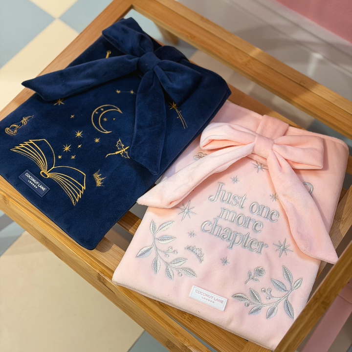 Book Sleeve made from soft blush pink velvet with grey embroidered floral details, stars and 'Just One More Chapter' lettering, finished with a large pink velvet bow and Coconut Lane logo, 