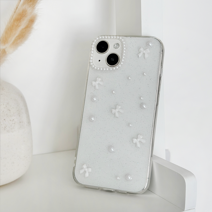A white smartphone with a Mini White Bows Phone Case by Coconut Lane, featuring small 3D flowers and pearl-like accents, is propped up against a white wall. In the background, a portion of a white vase with dried beige grass sticking out adds to the minimalist and monochromatic setting.