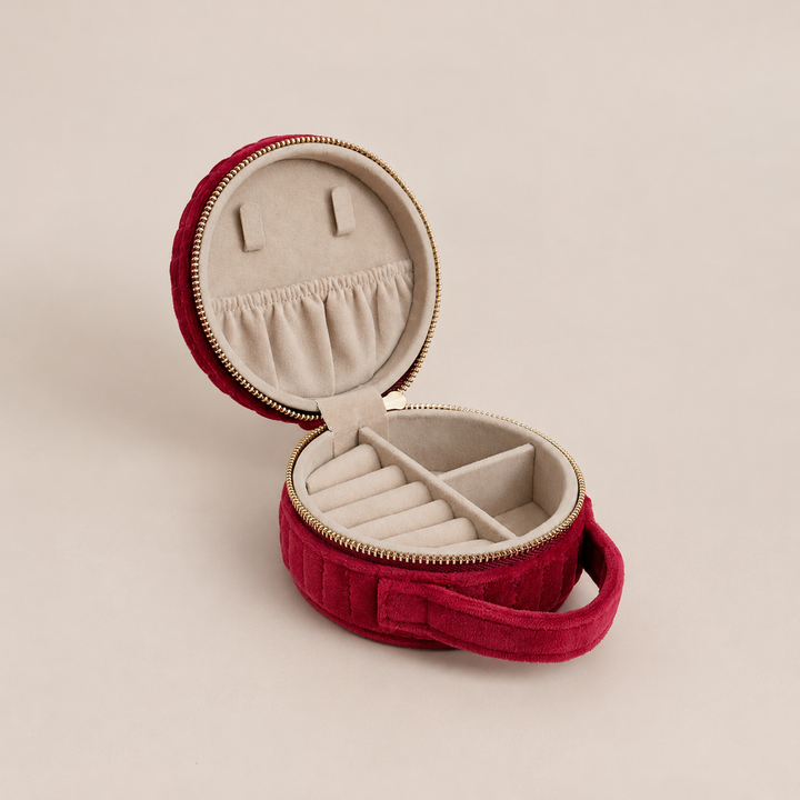 An open round red velvet jewelry box with a gold zipper, beige fabric lining, and internal compartments, set against a solid beige background.