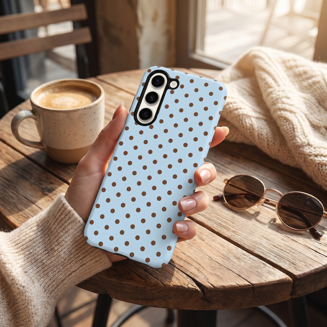 Person holding a phone with a blue polka dot case in a cosy setting with a cup of coffee and sunglasses.