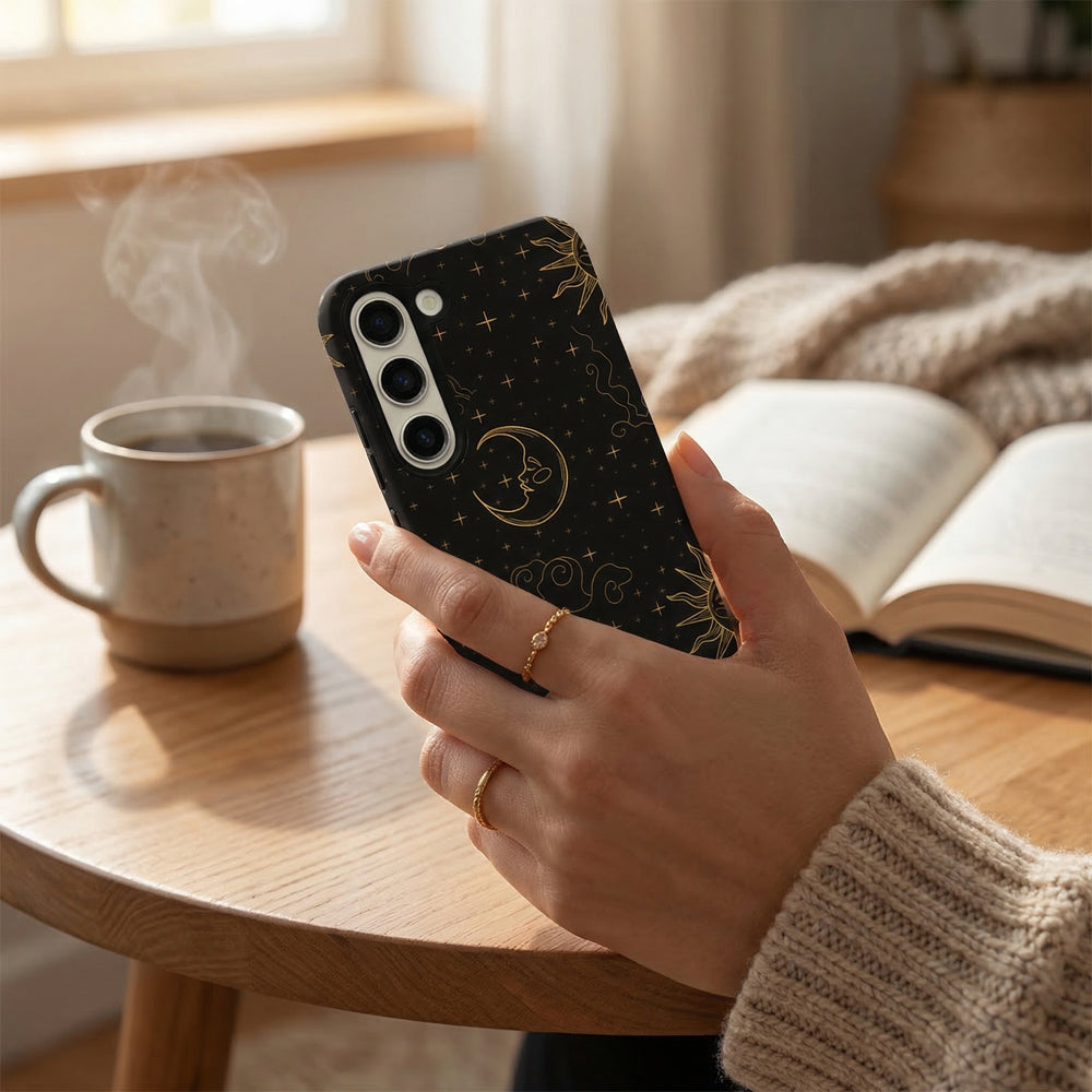 Hand holding a navy blue celestial-patterned Samsung phone case near an open book and steaming mug on a wooden table.
