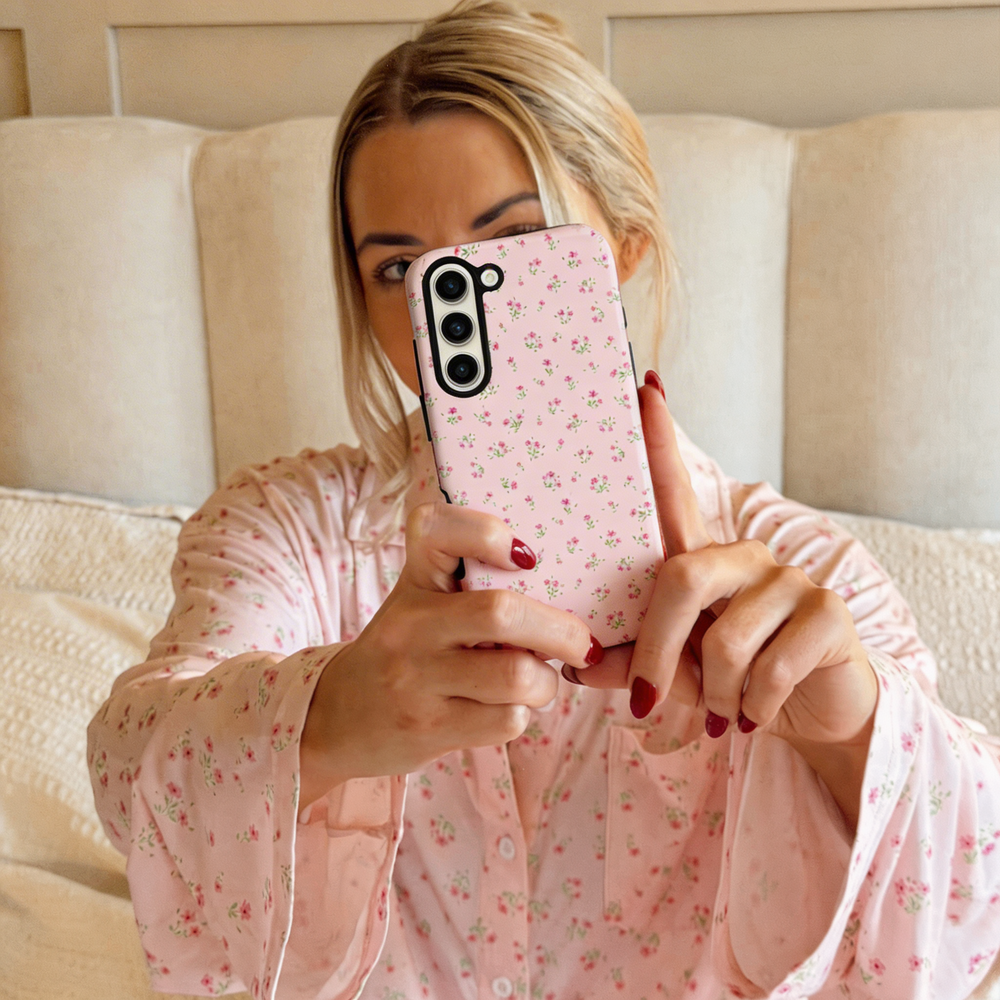 Woman in pink floral pajamas holds a Samsung phone with a pink ditsy floral Coconut Lane tough case on her bed.