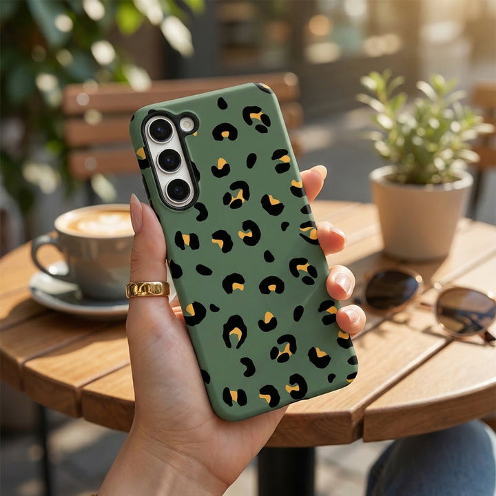 Hand holding a khaki leopard print Samsung Tough Phone Case at an outdoor café table with coffee and sunglasses.